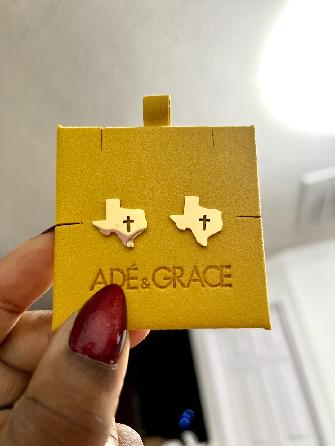 Gold Texas-shaped earrings with a cross in a 'Adé & Grace' box held by a hand with red nail polish.